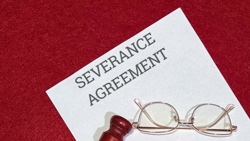 Severance agreement paperwork rests diagonally on a deep red textured surface beside reading glasses and a small wooden stamp representing final professional separation and legal formalities.